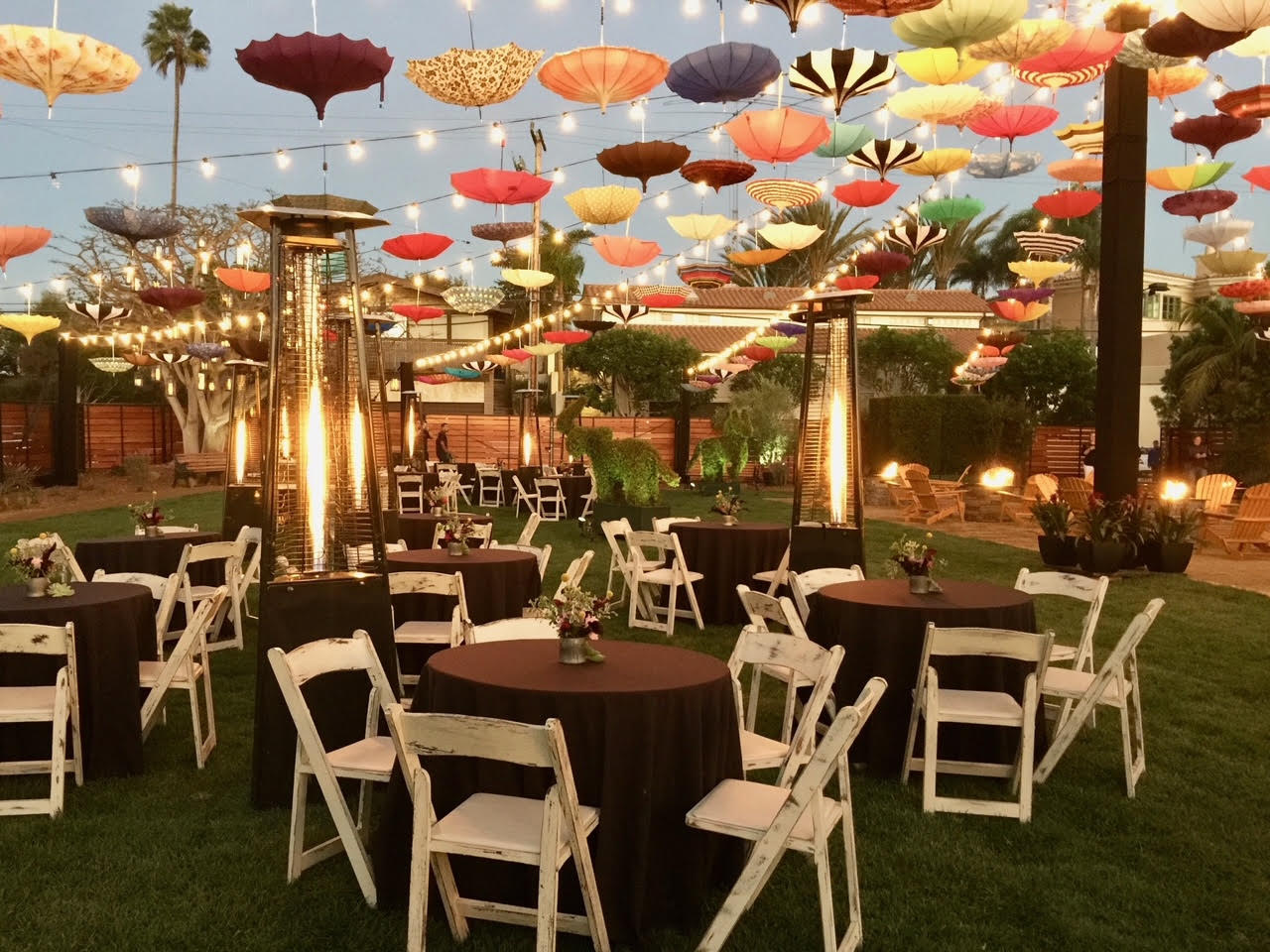 Outdoor event setup with tables and chairs set up underneath strands of large lights and upside-down hanging umbrellas